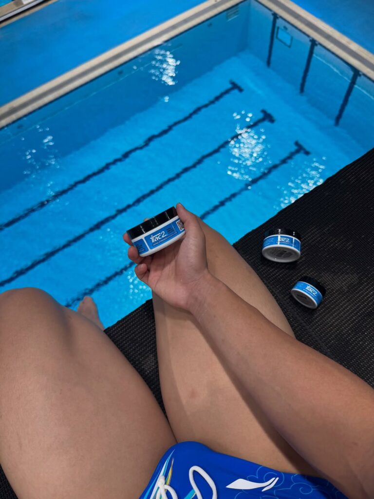 Person sitting by a blue swimming pool, holding a small blue-and-white tub with two more containers on a black lounge mat nearby.