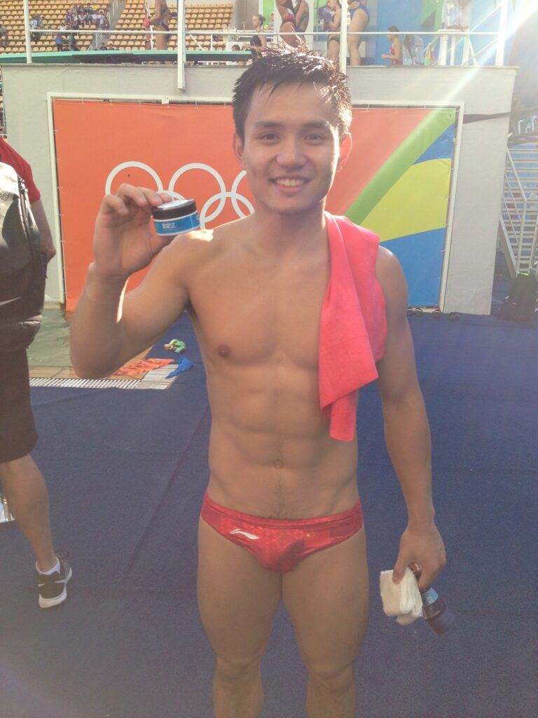 Smiling male swimmer shirtless, red briefs, pink towel over shoulder, holding a small blue jar at a sports venue with orange Olympic banner in background