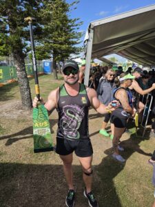 Man in black and green athletic outfit holding a green and yellow dragon-patterned board at a crowded outdoor race event under a canopy, facing the camera with a smile.