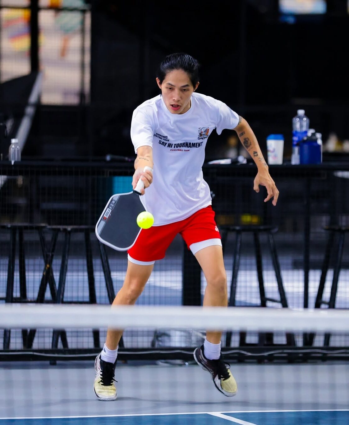 Male player in a white t-shirt and red shorts lunges to hit a bright yellow ball with a paddle on an indoor court.