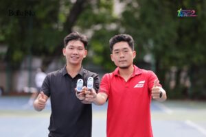 Two men stand outdoors on a sports court, smiling and giving thumbs up while displaying small bottles to the camera; logos for Belle Isle Sport and iTac2 appear in the background left and right.