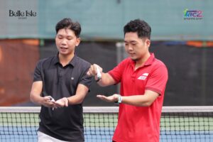 Two men on a tennis/paddle court near the net, one in black shirt and one in red polo, reaching as if about to hit a ball; logos in the background.