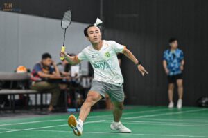 Male badminton player in a white jersey lunges forward on a green indoor court, swinging his racket mid-air as a shuttlecock approaches above him. Background includes blurred spectators and a dark wall.