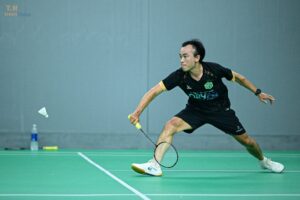 Male badminton player in a black shirt lunges to hit the shuttlecock on a green court court side view.