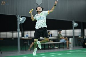 Male badminton player in mid-air, racket raised, about to smash on an indoor court; white jersey with sponsor logo, neon green shoals