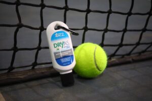 A bottle of DRYAS antiperspirant powder clipped to a tennis racquet grip with a bright tennis ball resting nearby against a tennis net backdrop.