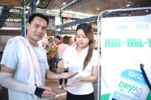 Man in a light blue shirt with a towel over his shoulder stands at a booth while a woman in white notes on a pad beside a DRYAS display sign.