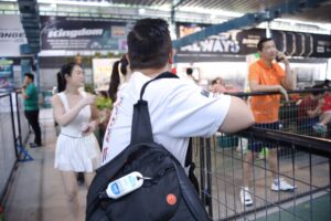 Man in a white shirt with a black backpack faces away, standing near a railing at an indoor event with others in the background.